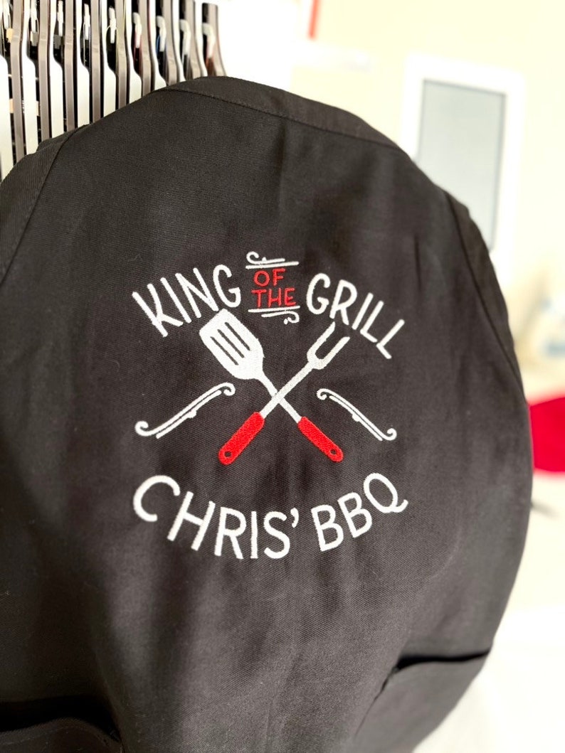 Pode incluir: Avental preto com texto bordado branco e um design de esp&aacute;tula e garfo cruzados. O texto diz "KING OF THE GRILL" acima de "CHRIS' BBQ". A esp&aacute;tula e o garfo t&ecirc;m cabos vermelhos. O avental est&aacute; pendurado.