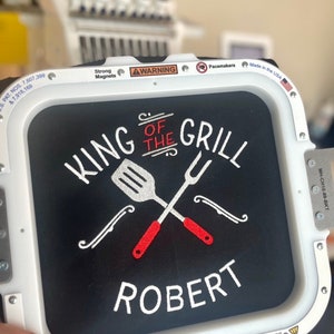 Pode incluir: Tecido preto bordado com "KING OF THE GRILL" e o nome "ROBERT" em branco. Uma esp&aacute;tula e um garfo s&atilde;o cruzados acima do nome, com detalhes em vermelho. O bordado est&aacute; em uma moldura branca, com avisos e o texto "Made in the USA".
