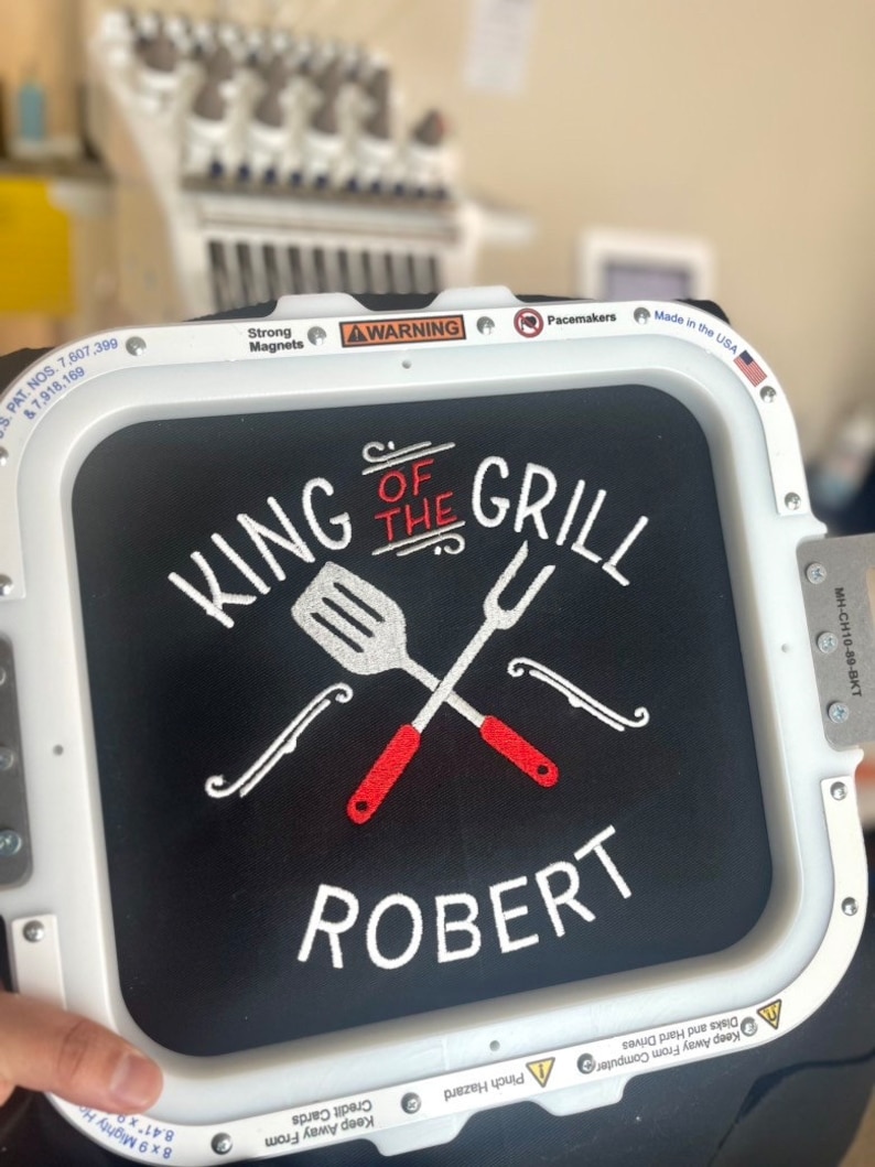 Pode incluir: Tecido preto bordado com "KING OF THE GRILL" e o nome "ROBERT" em branco. Uma esp&aacute;tula e um garfo s&atilde;o cruzados acima do nome, com detalhes em vermelho. O bordado est&aacute; em uma moldura branca, com avisos e o texto "Made in the USA".