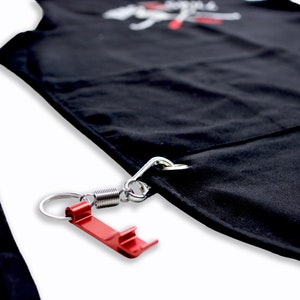 Pode incluir: Avental preto com um mosquet&atilde;o de mola de metal prateado e um abridor de garrafas vermelho preso. O avental tem um bolso e uma al&ccedil;a. O avental tem texto branco no topo.