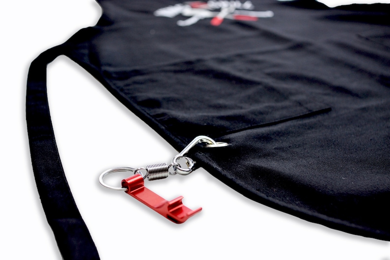 Pode incluir: Avental preto com um mosquet&atilde;o de mola de metal prateado e um abridor de garrafas vermelho preso. O avental tem um bolso e uma al&ccedil;a. O avental tem texto branco no topo.