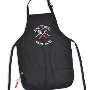 Pode incluir: Avental preto com dois bolsos frontais e uma al&ccedil;a de pesco&ccedil;o. O avental apresenta o texto "KING OF THE GRILL" e "NAME HERE" em branco, juntamente com utens&iacute;lios de grelhar vermelhos e brancos cruzados. Um chaveiro vermelho e prateado est&aacute; preso.