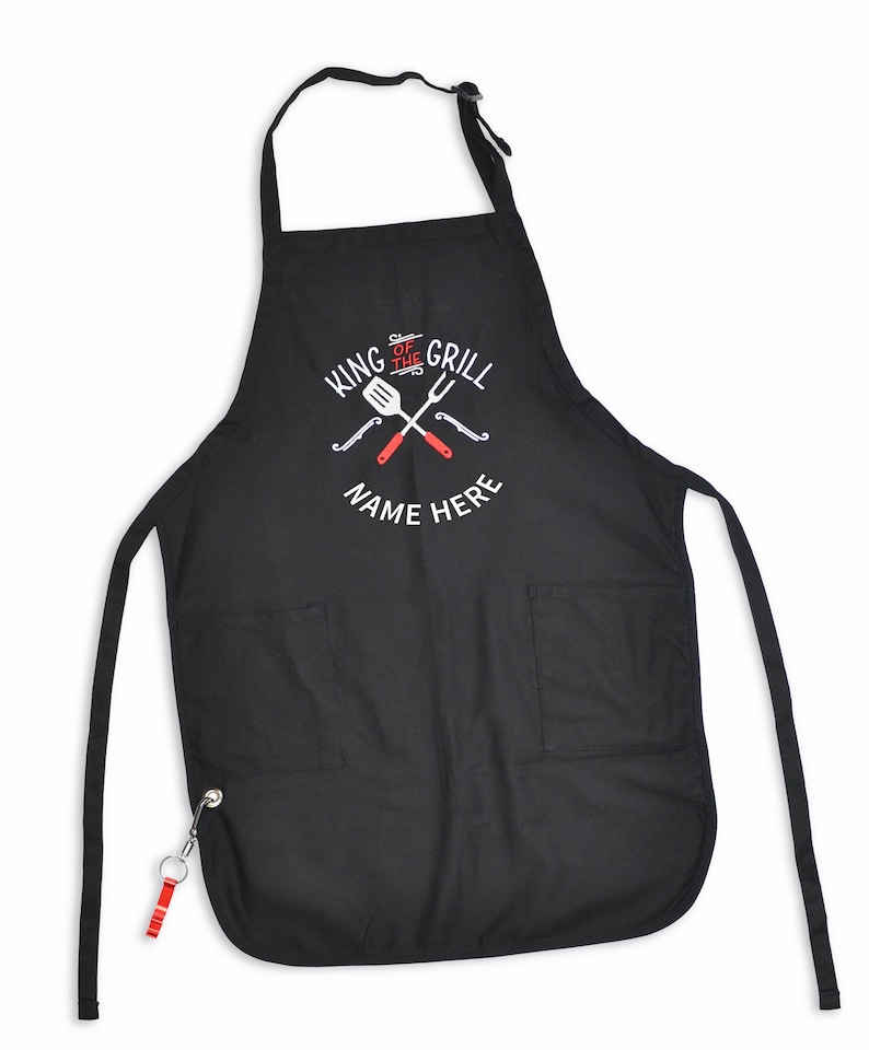 Pode incluir: Avental preto com dois bolsos frontais e uma al&ccedil;a de pesco&ccedil;o. O avental apresenta o texto "KING OF THE GRILL" e "NAME HERE" em branco, juntamente com utens&iacute;lios de grelhar vermelhos e brancos cruzados. Um chaveiro vermelho e prateado est&aacute; preso.