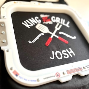 Pode incluir: Avental preto com texto bordado branco que diz "KING OF THE GRILL" com um design de esp&aacute;tula e garfo cruzados. O nome "JOSH" &eacute; bordado abaixo. O avental tem uma borda branca.