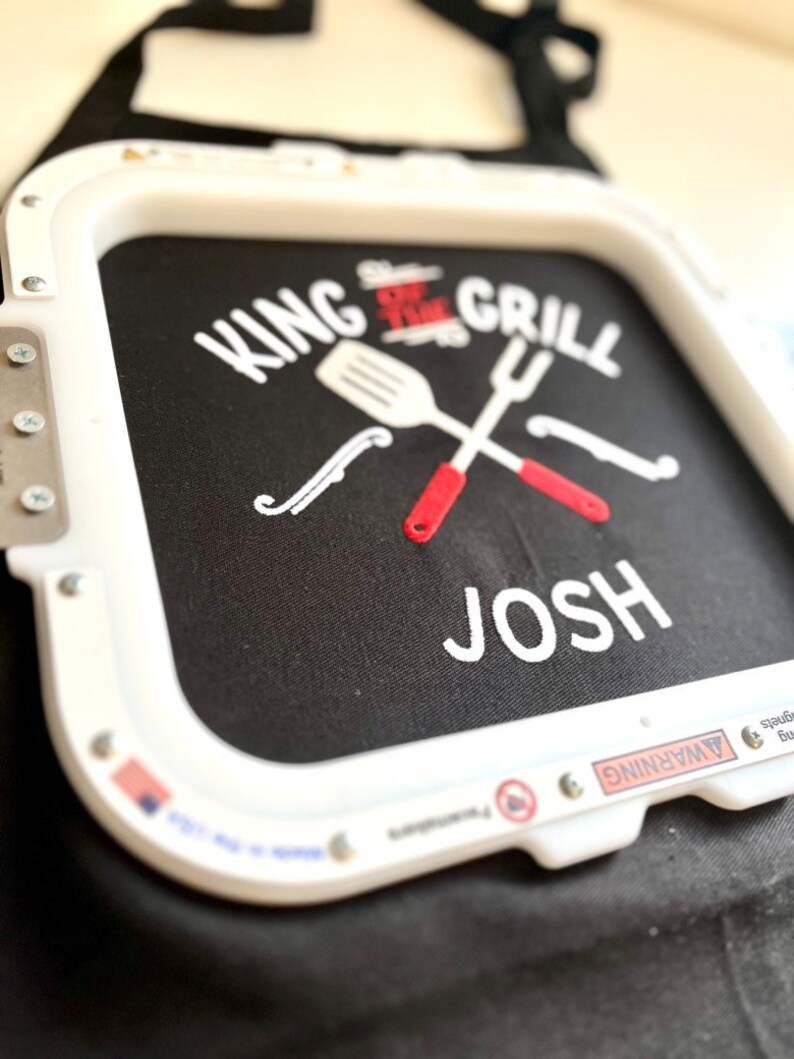 Pode incluir: Avental preto com texto bordado branco que diz "KING OF THE GRILL" com um design de esp&aacute;tula e garfo cruzados. O nome "JOSH" &eacute; bordado abaixo. O avental tem uma borda branca.