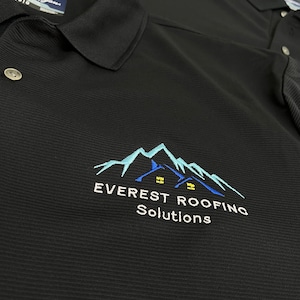 May include: Black polo shirt with a white embroidered logo that says "EVEREST ROOFING Solutions" and features a blue mountain range with two houses.