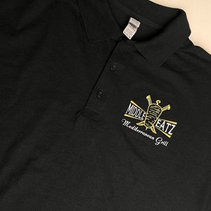 May include: Black polo shirt with a gold embroidered logo that says "Middle EatZ Mediterranean Grill".