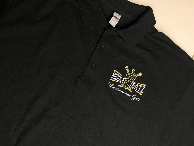 May include: Black polo shirt with a gold embroidered logo that says "Middle EatZ Mediterranean Grill".