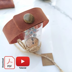 May include: A brown leather bracelet with a glass vial containing dried flowers. The vial is sealed with a cork and tied with twine. The bracelet has a brass button closure.