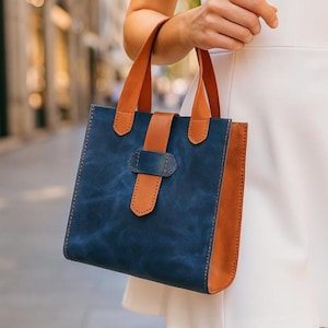 Puede incluir: Un bolso tote de cuero azul y marrón. El bolso tiene forma rectangular con asas y paneles laterales de cuero marrón, y una parte delantera y trasera de cuero azul. Una correa de cuero marrón asegura el bolso con una hebilla. El bolso es sostenido por una persona.