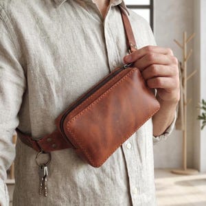 May include: A rectangular brown leather crossbody bag with a top zip closure and a matching leather strap. The bag features a metal ring attached to the side, holding a set of silver-toned keys. The leather has a smooth, matte finish.