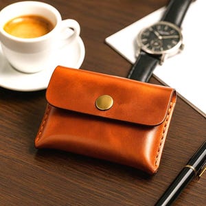 Op de afbeelding: Een bruine leren portemonnee met een gouden knoopsluiting. De portemonnee is rechthoekig met een gebogen flap. Een witte kop koffie, een horloge en een pen zijn ook zichtbaar.