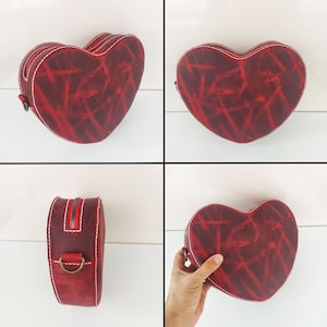 May include: Red leather heart-shaped handbag with a zipper closure and a strap. The bag has a distressed look with a red and white pattern.