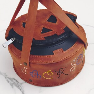 May include: A brown leather pumpkin-shaped bag with a black zipper and a black leather lining. The bag has a top handle and is embroidered with the word "jack-o-lantern" in colorful thread.
