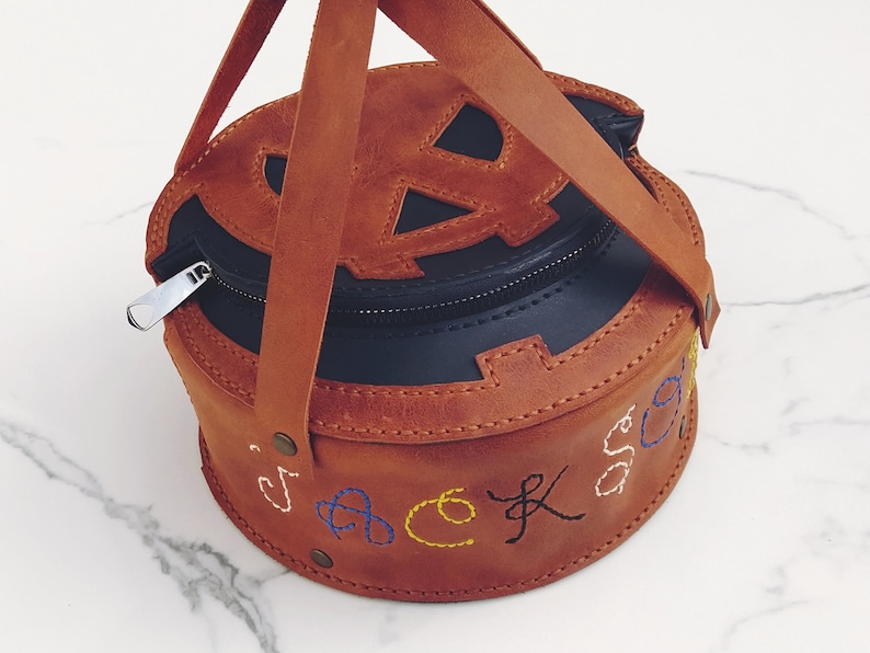 May include: A brown leather pumpkin-shaped bag with a black zipper and a black leather lining. The bag has a top handle and is embroidered with the word "jack-o-lantern" in colorful thread.