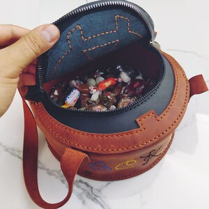 May include: A brown leather round bag with a zipper closure and a strap. The bag is open and filled with colorful candies.