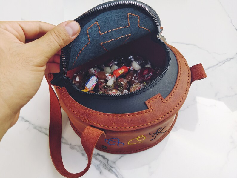 May include: A brown leather round bag with a zipper closure and a strap. The bag is open and filled with colorful candies.