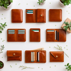 May include: A collection of various brown leather wallets, including cardholders, bi-fold wallets, and coin purses. Some wallets are open, displaying cards and cash. The wallets are arranged on a white surface with small green plants and a gift box.