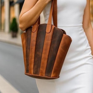 May include: A brown and tan leather tote bag with vertical panels. The bag has two handles and a unique, fan-like shape. The leather has a distressed appearance, adding to its rustic charm. The bag is carried by a person wearing a white dress.