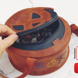May include: A brown leather round bag with a zipper closure and a black interior. The bag has a stitched design on the front and a strap. The bag is open and filled with colorful stones.