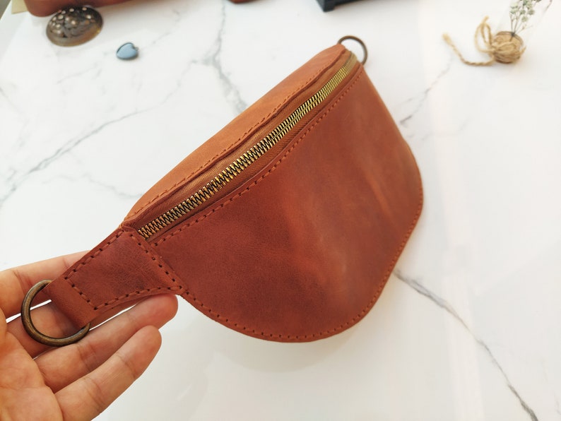 Pode incluir: Uma pochete de couro marrom com um z&iacute;per dourado e uma fivela de metal. A bolsa &eacute; feita de couro macio e flex&iacute;vel e tem um design cl&aacute;ssico e atemporal.