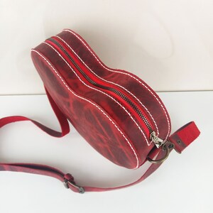 May include: A red leather handbag with a heart shape and a white stitch detail. The bag has a zipper closure and a long adjustable strap.