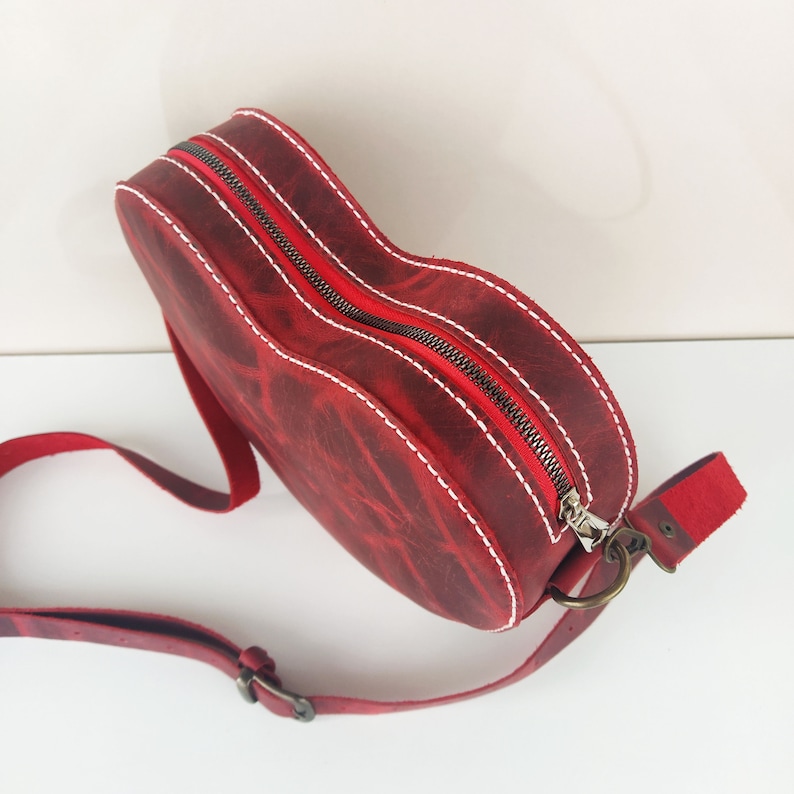 May include: A red leather handbag with a heart shape and a white stitch detail. The bag has a zipper closure and a long adjustable strap.