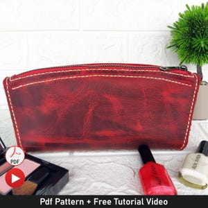 Puede incluir: Bolsa de cuero roja con cremallera y costuras blancas. La bolsa está sobre un fondo de ladrillos blancos con botellas de esmalte de uñas y pinceles de maquillaje.