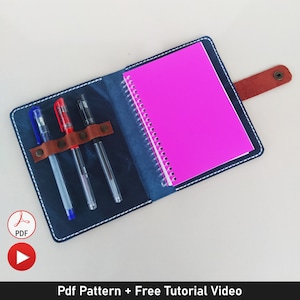 May include: A blue leather notebook cover with a pink spiral notebook inside. The cover has a flap with a brown leather strap and a metal snap closure. There are three pens in the pen slots, one red and two blue.