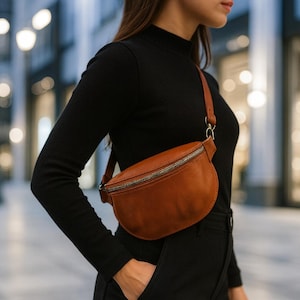 Puede incluir: Un bolso de cintura de cuero marrón con cierre de cremallera plateada. La bolsa se lleva cruzada con una correa marrón. El bolso de cintura tiene forma de media luna y parece estar hecho de cuero suave.