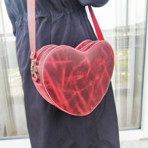 May include: A red leather heart-shaped crossbody bag with a white stitch detail and a gold-toned zipper.