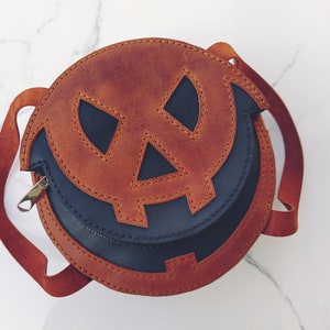 May include: A brown leather pumpkin-shaped crossbody bag with a black leather lining. The bag has a stitched jack-o'-lantern face with a stitched smile.