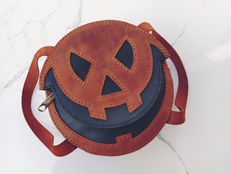May include: A brown leather pumpkin-shaped crossbody bag with a black leather lining. The bag has a stitched jack-o'-lantern face with a stitched smile.