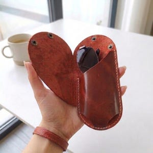 May include: A heart-shaped brown leather glasses case with visible stitching along the edges and two metal snap fasteners at the top. The case is partially open, revealing a pair of dark-tinted sunglasses tucked inside the structured leather pouch.