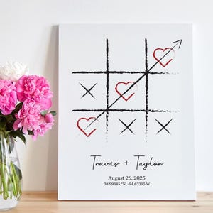 May include: A white canvas print featuring a tic-tac-toe design with hearts and crosses. The names "Travis + Taylor" and the date August 26, 2025, are printed below. A vase of pink and white peonies sits to the left.