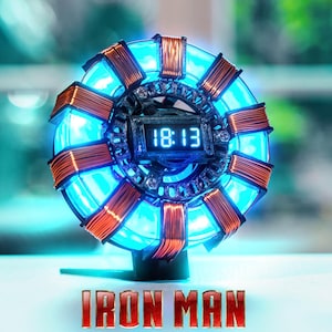 Può includere: Un orologio circolare in stile reattore ad arco, con un design blu e rame. Il display digitale mostra l'ora 18:13. Le parole "IRON MAN" sono visualizzate in rosso nella parte inferiore.