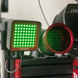 May include: A black and red electronic device with the word "REDION" printed on it. The device has a grid of green lights and is connected to a black cable. A red and green cylindrical component is also visible.