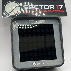 May include: A gray electronic device with a carbon fiber pattern frame and a black screen. The device has the text "SECTOR 17" and "RACING ENGINEERING" on a black label above it. The device also has the "SECTOR 17" logo on the bottom.