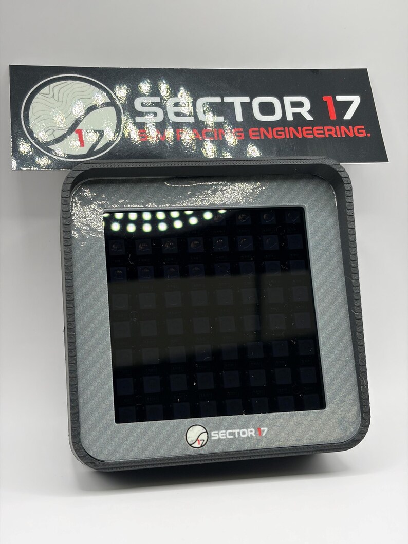 May include: A gray electronic device with a carbon fiber pattern frame and a black screen. The device has the text "SECTOR 17" and "RACING ENGINEERING" on a black label above it. The device also has the "SECTOR 17" logo on the bottom.