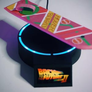 Puede incluir: Un aeropatín rosa, amarillo y verde de la película Regreso al Futuro II, apoyado sobre una base negra con un anillo azul brillante. La base tiene el logotipo de la película.