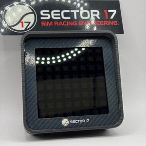 May include: A black and gray digital display device with the text "SECTOR 17 SIM RACING ENGINEERING." The device has a carbon fiber texture around the screen and the logo "SECTOR 17" at the bottom.