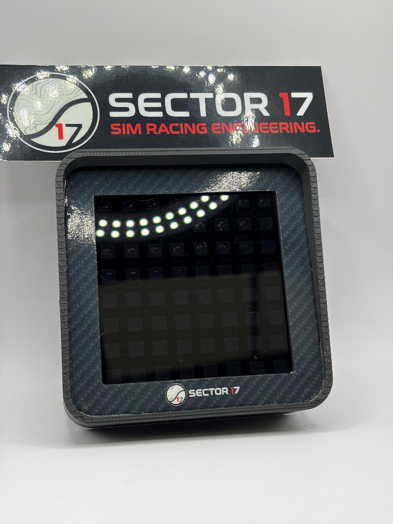 May include: A black and gray digital display device with the text "SECTOR 17 SIM RACING ENGINEERING." The device has a carbon fiber texture around the screen and the logo "SECTOR 17" at the bottom.