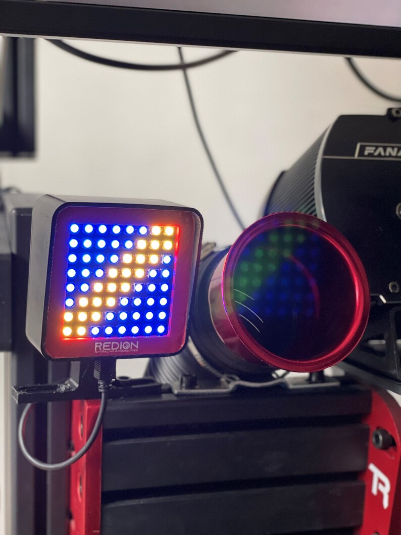 May include: A black and red electronic device with a grid of blue and yellow lights, displaying a pattern. The device is labeled "REDION" and is mounted on a black and red structure. A red-rimmed lens is visible.