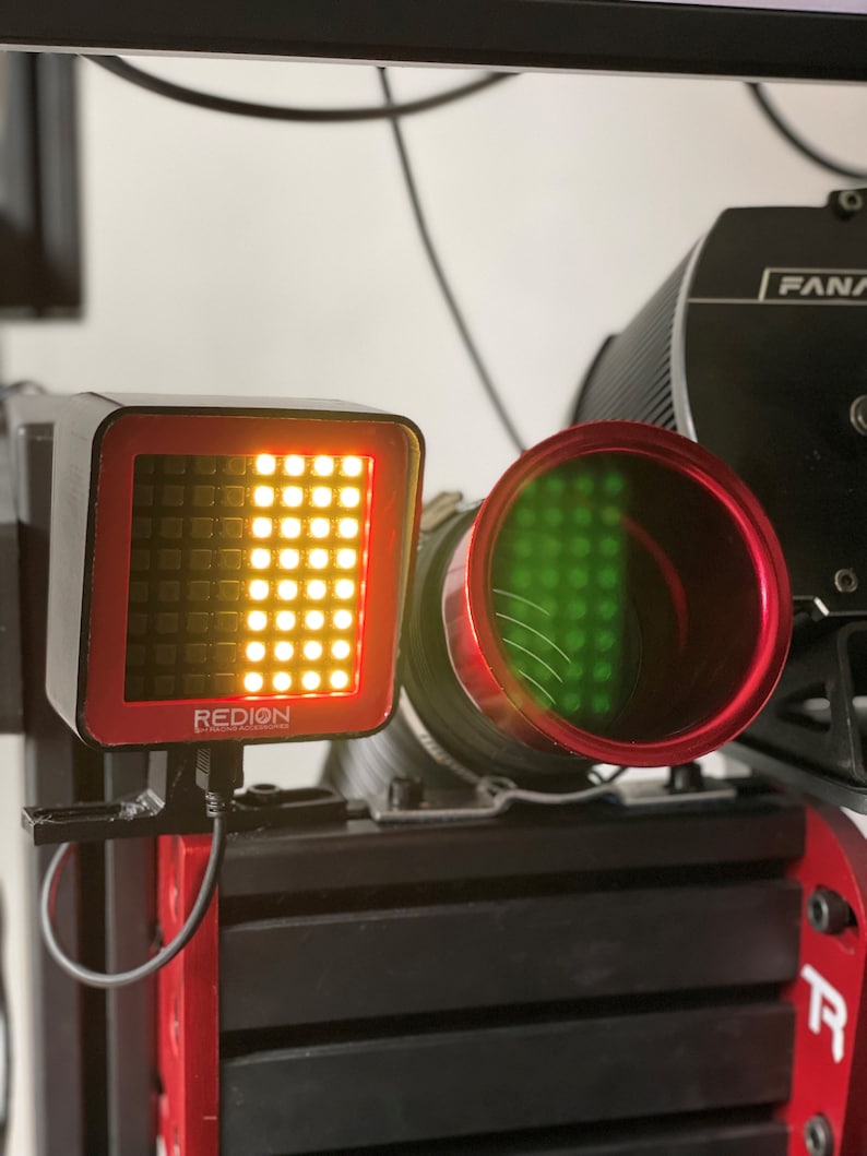 May include: A close-up of a racing simulator component. A square, black device with a grid of yellow lights and the word "REDION" is visible. A red-rimmed, cylindrical component with green lights is also present. The components are mounted on a black and red frame.