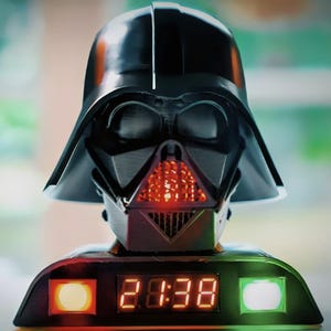 Könnte beinhalten: Schwarzer Wecker in Form eines Darth-Vader-Helms. Die Uhr zeigt die Zeit in orangefarbenen Digitalziffern an. Der Sockel hat orange und grüne Lichter auf beiden Seiten der Anzeige. Ein Sammlerstück zum Thema Star Wars.