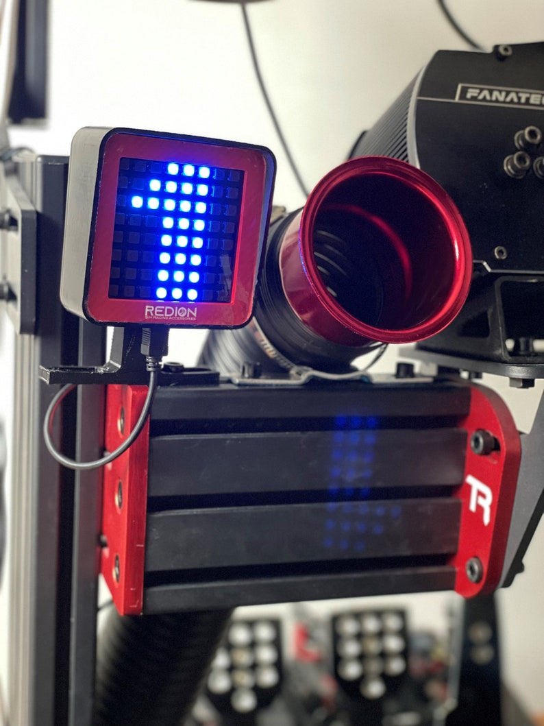 May include: A racing simulator setup featuring a red and black digital display with the word "REDION" and a blue arrow. The setup includes a red-rimmed component and a black structure with the "R" logo.
