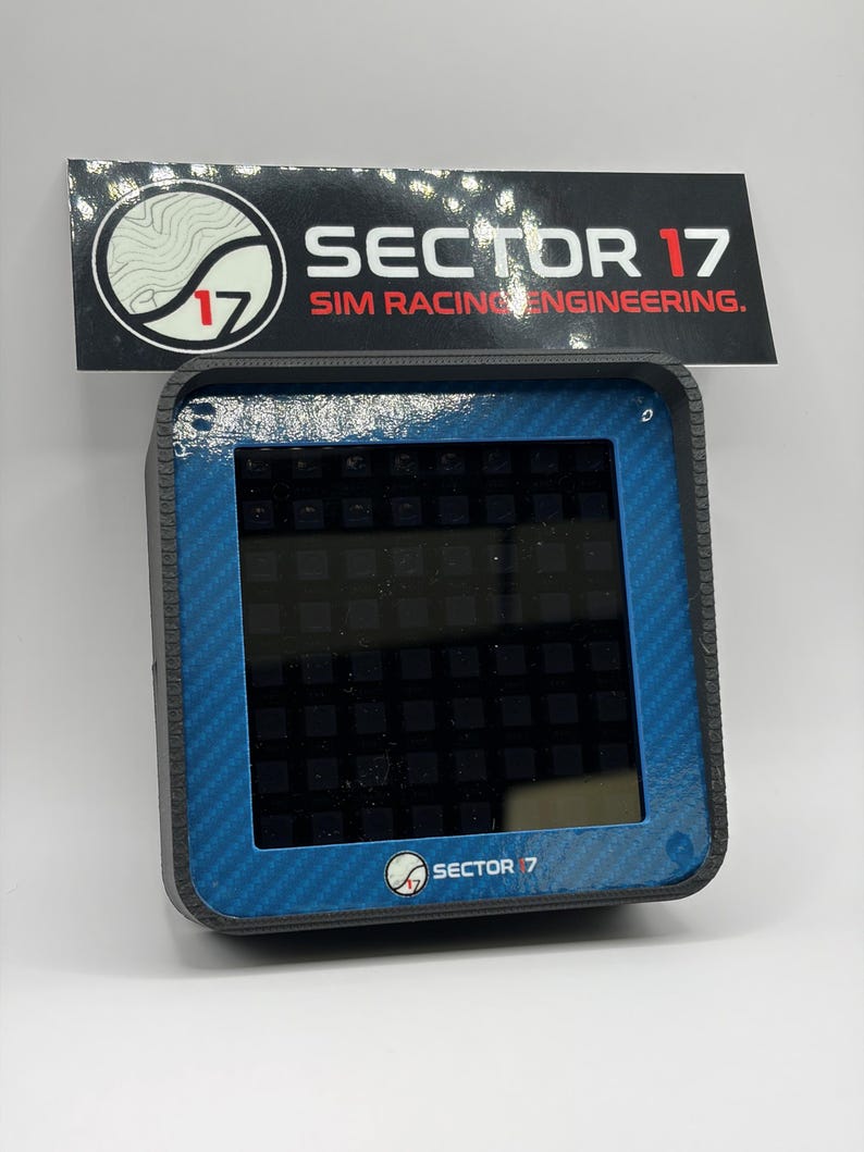May include: A black and blue electronic device with a screen, featuring the text "SECTOR 17 SIM RACING ENGINEERING." The device has a carbon fiber texture on the edges and the logo "SECTOR 7" at the bottom. The screen displays a grid pattern.