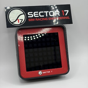 May include: A black and red electronic device with a screen, featuring the text "SECTOR 17 SIM RACING ENGINEERING." The device has a square screen with a grid pattern and a series of lights at the top. The logo of "SECTOR 17" is also visible.