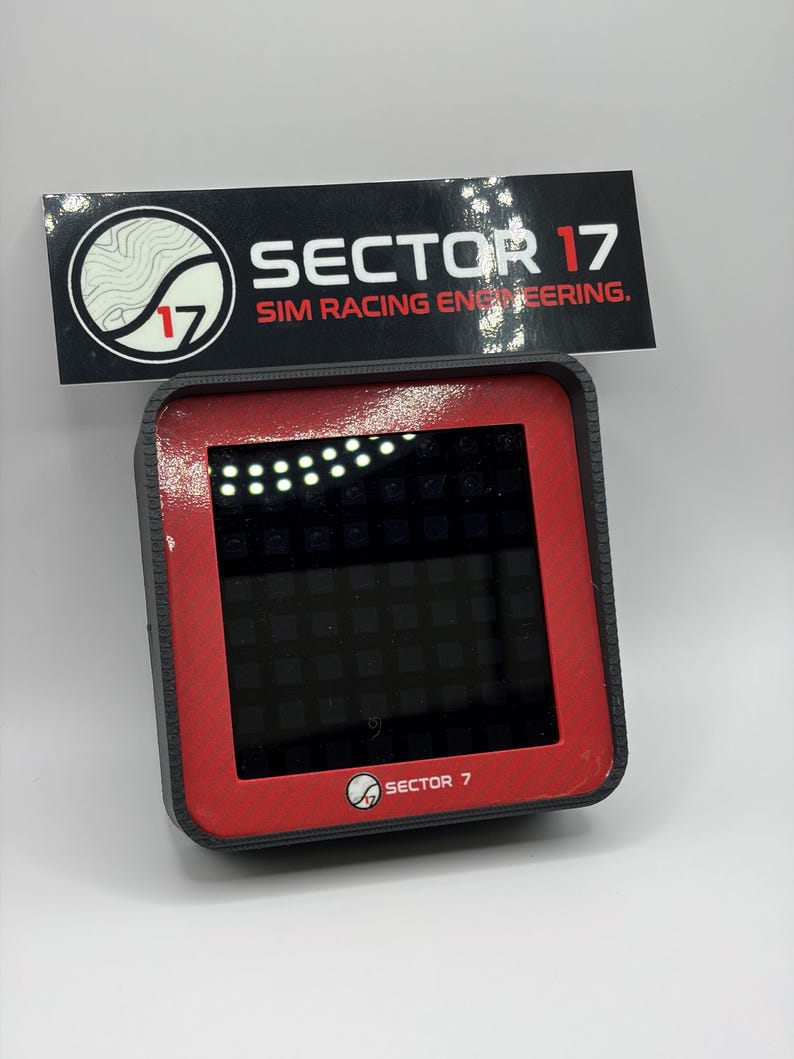 May include: A black and red electronic device with a screen, featuring the text "SECTOR 17 SIM RACING ENGINEERING." The device has a square screen with a grid pattern and a series of lights at the top. The logo of "SECTOR 17" is also visible.