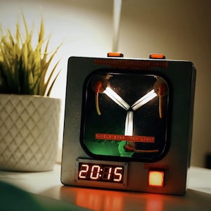 May include: A silver-coloured clock with a digital display showing "20:15". The clock has a unique design with glowing elements and a switch. A potted plant with green and yellow grass sits to the left.